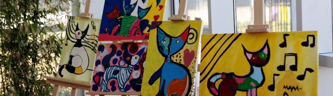 Colorful paintings of cats on easels in a bright room next to a plant