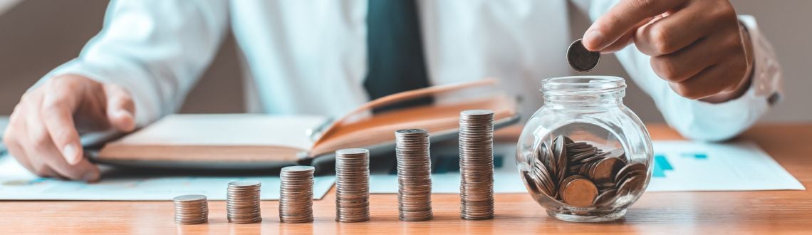 Stacks of coins going from small to large on a desk. A man in a tie is dropping a coin into a glass jar full of more coins