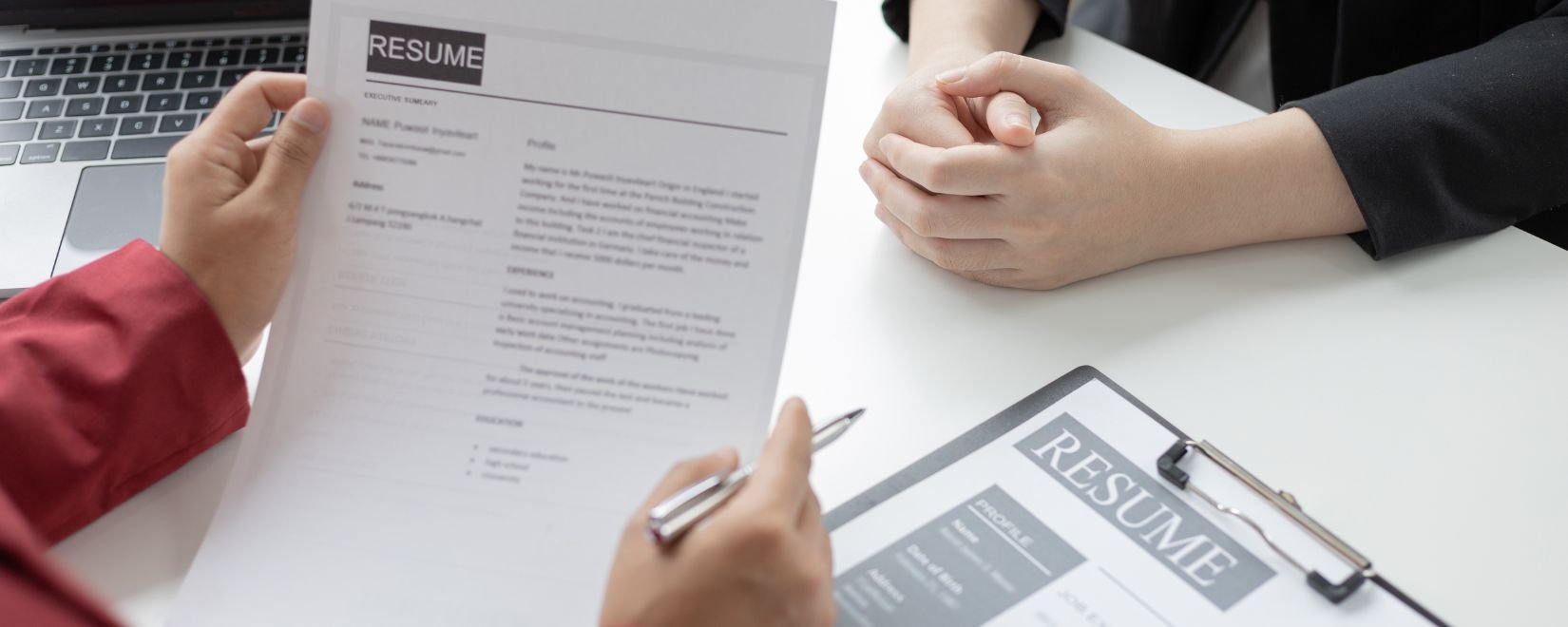 Close up of a hand holding a pen and a resume, with someone else's clasped hands across the desk