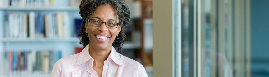 An African-American female librarian wearing glasses and a pink sweater, smiling in front of shelves of books
